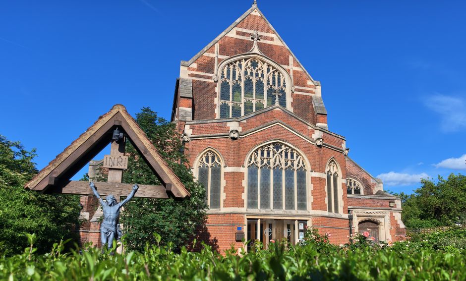 A view of a historic red-brick church with large stained glass windows and intricate stone tracery, set against a bright blue sky. In the foreground, a small wooden crucifix featuring a silver figure 
