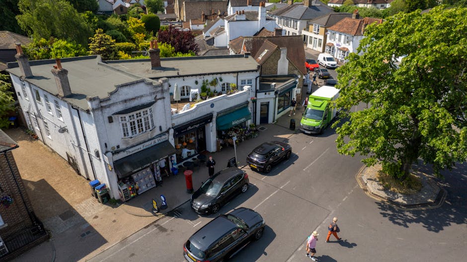 An aerial view of a small white building with green signage and awnings, situated on a busy street corner in Finchley. In front, there is a flower delivery shop with floral arrangements displayed outs