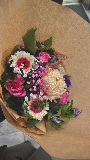 A vibrant floral bouquet featuring pink roses, white daisies with pink centers, a large cream-colored peony, and small purple and pink filler flowers, all arranged with lush green leaves. The flowers 